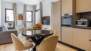 Ein modernes Wohnzimmer mit einem runden Tisch, beigen Sesseln, einem Flachbildfernseher und einer Küche mit Mikrowelle und Backofen.