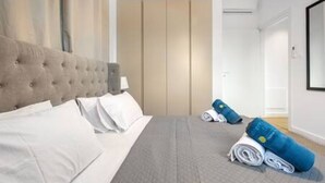 Un dormitorio con un cabecero capitonado, una cama con ropa de cama blanca, un edredón gris y toallas azules enrolladas en el suelo.