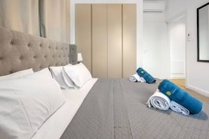 Ein Schlafzimmer mit einem gebürsteten Kopfende, einem Bett mit weißen Leinentüchern, einer grauen Decke und gerollten blauen Handtüchern auf dem Boden.