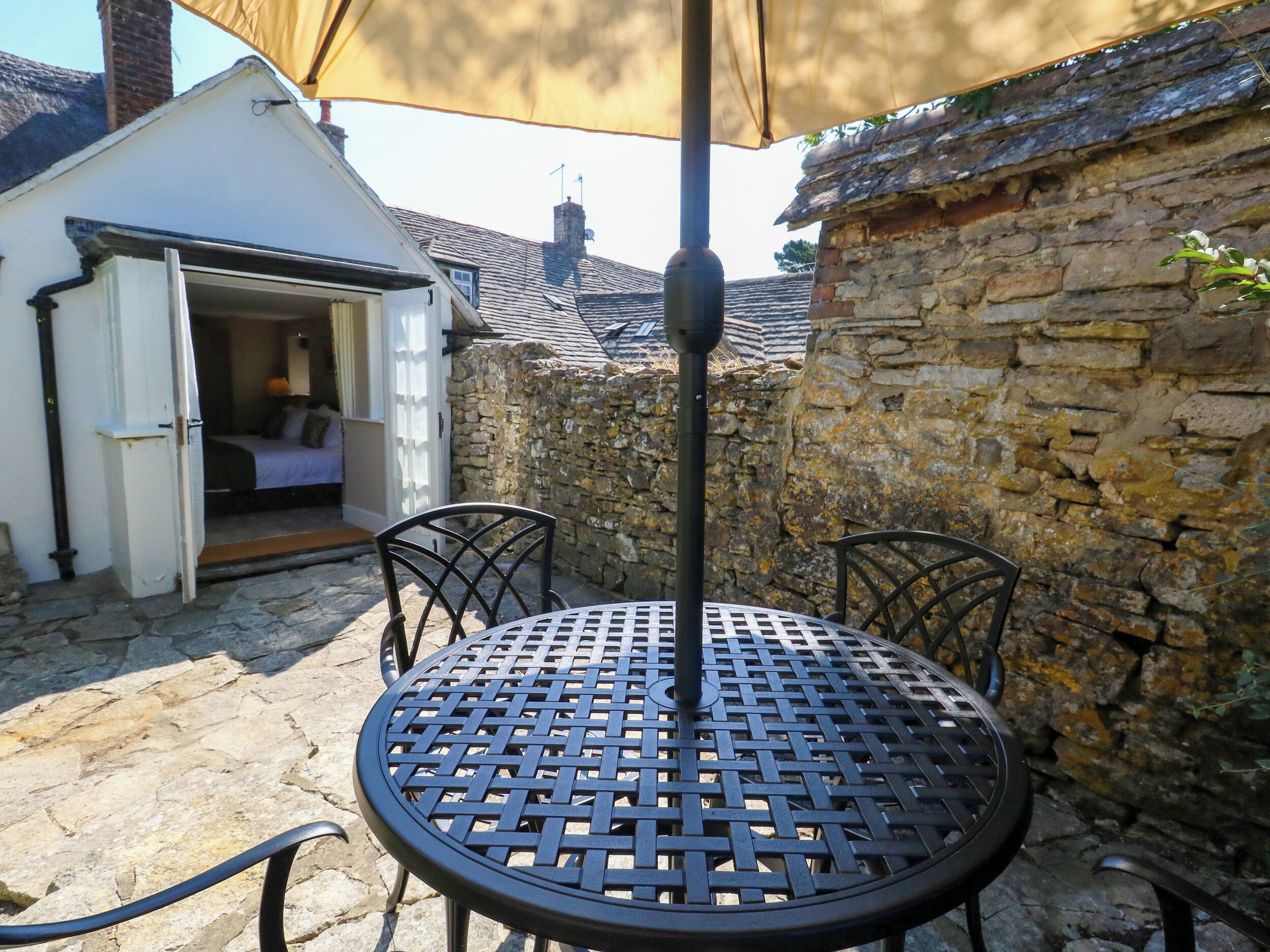 Une table ronde en métal avec un siège en osier, une petite pièce ouverte avec un lit et un mur en pierre.