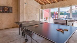 Ein Tischtennistisch mit Schläger und Bällen, eine Dartscheibe und ein Kicker befinden sich in einem Raum mit Betonboden und Blick ins Freie.