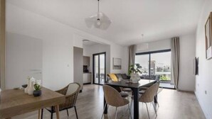 A modern dining area with a wooden table and chairs, a hanging light fixture, and a view through large windows. - 202 Piliou Large Balcony & Shared Rooftop Pool (Limassol)