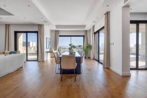 A spacious living area with a dining table, a sofa, and large windows offering a city view. - 301 Piliou - Large Terrace & Rooftop Communal Pool (Limassol)