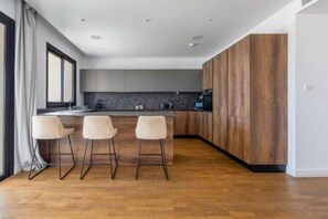 A modern kitchen with wooden cabinets, a dark countertop, and three bar stools. - 301 Piliou - Large Terrace & Rooftop Communal Pool (Limassol)