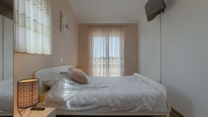 Een slaapkamer met een bed, een bedlampje, een raam met gordijnen en een aan de muur bevestigde televisie.