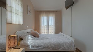 Ein Schlafzimmer mit einem Bett, einer Nachttischlampe, einem Fenster mit Vorhängen und einem an der Wand montierten Fernseher.