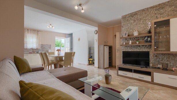 Een moderne woonkamer met een bank, salontafel en een flatscreen televisie. Er is een eethoek met stoelen en een tafel, en een keuken met een koelkast en kastjes.