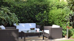Een terras buiten met rieten meubels, een glazen tafel en een witte bank.