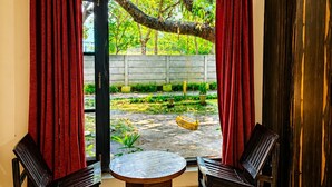 A room with a sliding glass door leading to a garden, two black chairs, a small round table, and a dark wooden cabinet. - Wildline Gold Resort (Ramnagar)