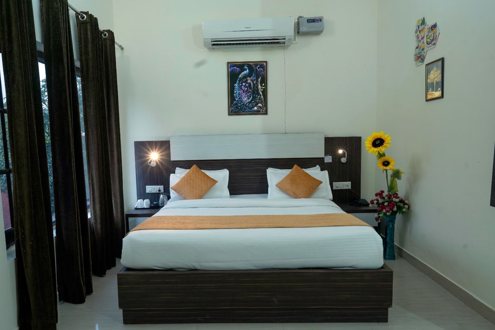Une chambre d’hôtel avec un grand lit, une tête de lit en bois foncé, du linge de lit blanc, une table de chevet avec une lampe, un climatiseur fixé au mur et un vase rempli de tournesols.