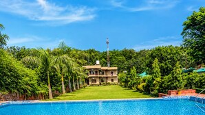 A swimming pool with a tiled edge, surrounded by lush greenery and a building in the background. - Wildline Gold Resort (Ramnagar)