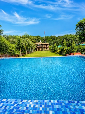 Una piscina con borde de azulejos, rodeada de exuberante vegetación y un edificio al fondo.