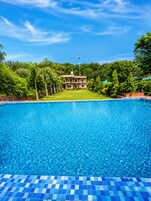 Una piscina con borde de azulejos, rodeada de exuberante vegetación y un edificio al fondo.