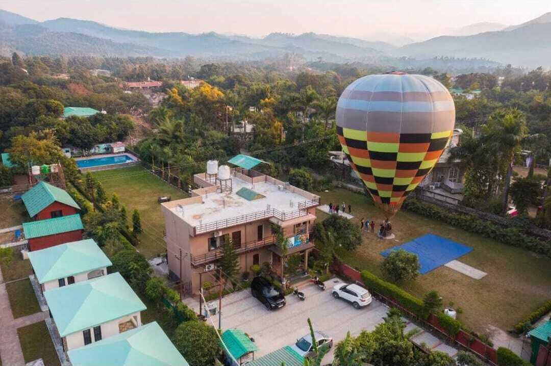 A hot air balloon over a residential area with houses, a swimming pool, and a large building with a flat roof.