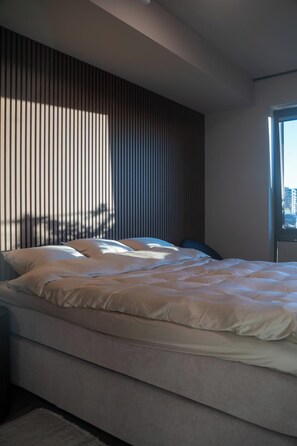 Une chambre moderne avec un grand lit, une tĂȘte de lit Ă lattes et une fenĂȘtre donnant sur la ville.