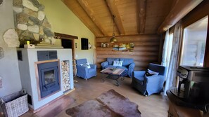 Een knus interieur van een chalet met een stenen open haard, blauwe gestoffeerde meubels, een houten plafond en een groot raam.