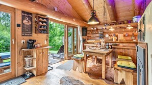 Interior d'una cabana de fusta amb zona de menjador, cuina i una finestra gran que dona a vegetació.