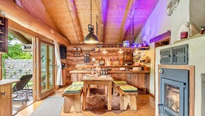 Interior d'una cabana de fusta amb taula de dinar, bancs, una llar de foc i zona de cuina amb prestatges i electrodomèstics.
