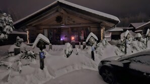 Un edificio cubierto de nieve con fachada de madera, una entrada visible y un área de estacionamiento con coches nevados.