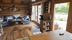 Een knus interieur van een blokhut met een bank, fauteuils, een houten eettafel en een keukenhoek met planken.