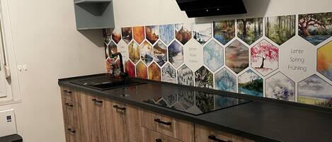 A kitchen with wooden cabinets, a black range hood, a washing machine, and a tiled backsplash with seasonal scenes.
