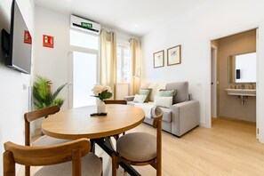 Ein Wohnzimmer mit einem runden Holztisch, zwei Stühlen, einer Couch, einem Fernseher und einem Fenster mit Vorhängen.