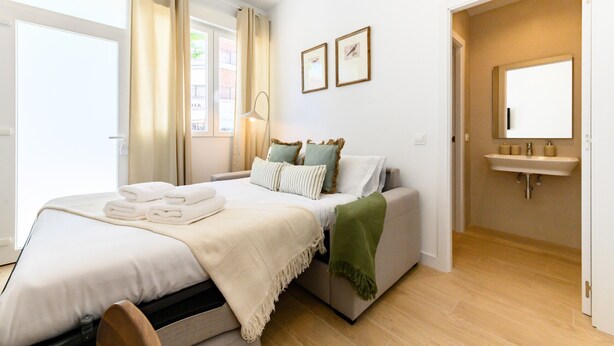 A hotel room with a bed, a bedside table, a lamp, a window with curtains, a wall-mounted sink, and a door leading to another room. - (SDB1L2) 1 bedroom apartment in Tetuán, Madrid (Madrid)