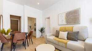 Un salon avec un canapé, une table basse et des fauteuils, un coin repas avec une table et des chaises, et une cuisine en arrière-plan.