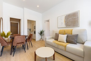 Ein Wohnzimmer mit einer Couch, Couchtisch und Sesseln, ein Essbereich mit Tisch und Stühlen sowie eine Küche im Hintergrund.
