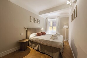 A bedroom with a bed, a bedside lamp, a window with curtains, and a small round table. - Emma by MMega (Firenze)