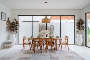 Un comedor con una mesa dispuesta para seis personas, rodeada de sillas de madera, una lámpara colgante y plantas en macetas grandes.