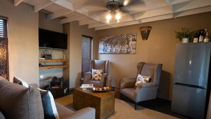 A living room with a sofa, two armchairs, a coffee table, a flat-screen TV, a ceiling fan, and a refrigerator.