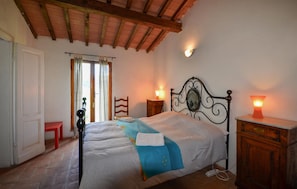Ein Schlafzimmer mit Bett, Holz-Kopfteil, Nachttischen, einem Beistelltisch, einem roten Hocker, einem Fenster mit Vorhängen und einer Holzbalkendecke.