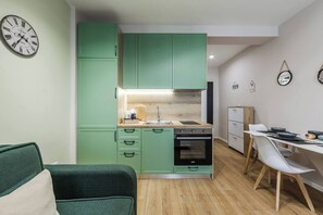 Una cocina compacta con gabinetes verdes, un fregadero y un horno. Hay un comedor con una mesa y sillas blancas.