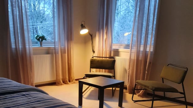 A hotel room with a bed, a table, two chairs, a lamp, and large windows with curtains. - Going North (Molkom)