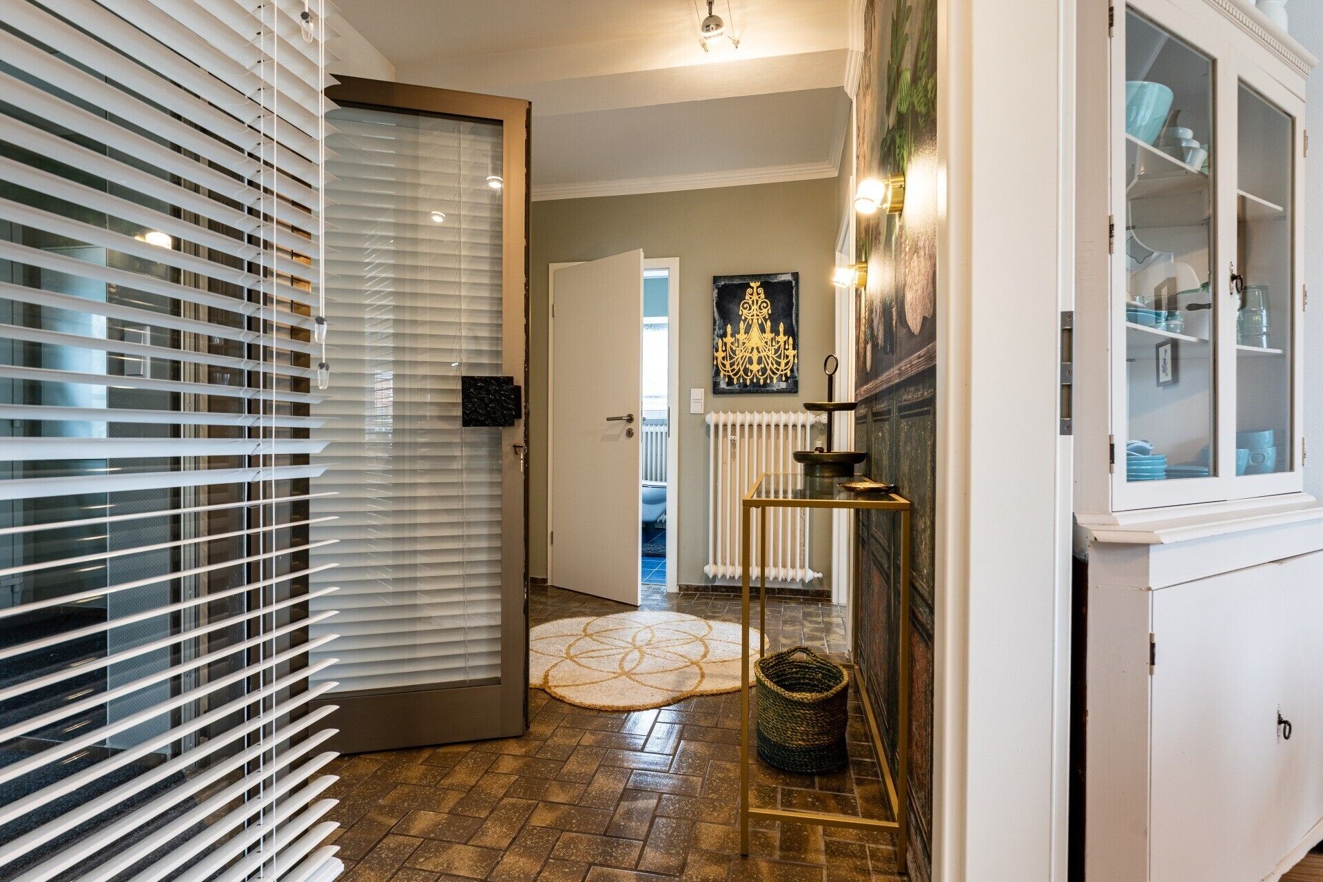 A hallway with a door, a radiator, a patterned rug, a small table with a candle, and a cabinet with glass doors.