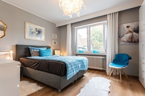 Une chambre à coucher avec un lit, une chaise, un radiateur, une fenêtre et des œuvres d’art accrochées aux murs.