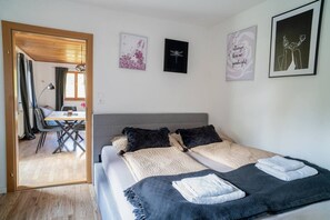A bedroom with a bed, pillows, a bedside table, a chair, and a desk. There are wall art pieces and a door leading to another room.