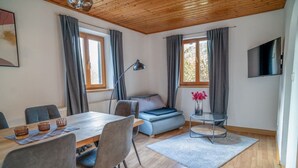 Habitació moderna amb sostre de fusta, taula de dinar amb cadires, sofà, taula rodona petita amb un ram i televisió penjada a la paret.