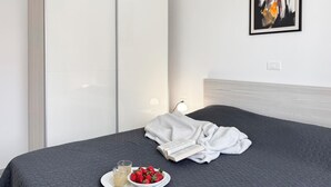 Uma cama bem arrumada com um edredom escuro, um roupão branco, um prato com morangos e um copo de bebida, e uma luminária na mesinha de cabeceira.