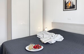 Ein ordentlich bezogenes Bett mit einer dunklen Tagesdecke, einem weißen Bademantel, einem Teller mit Erdbeeren und einem Glas Getränk, sowie eine Lampe auf dem Nachttisch.