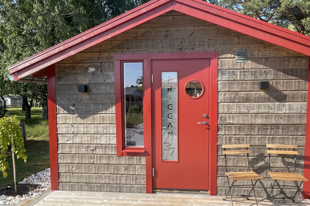 A small wooden cabin with a red door and a window, two foldable chairs outside, and a clear sky in the background.
