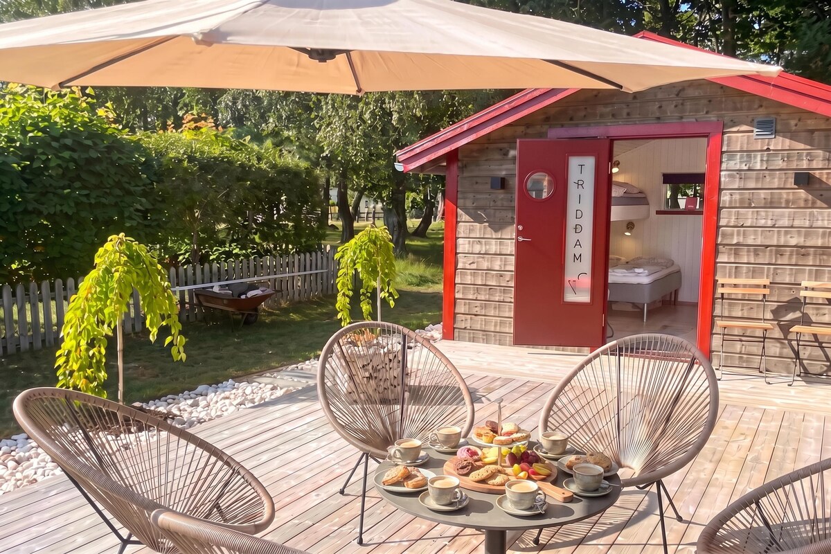 Eine Holzterrasse mit Korbstühlen, einem Tisch mit Kaffee und Gebäck und ein kleines rotes Gebäude mit dem Schild 'TRIDAM'.
