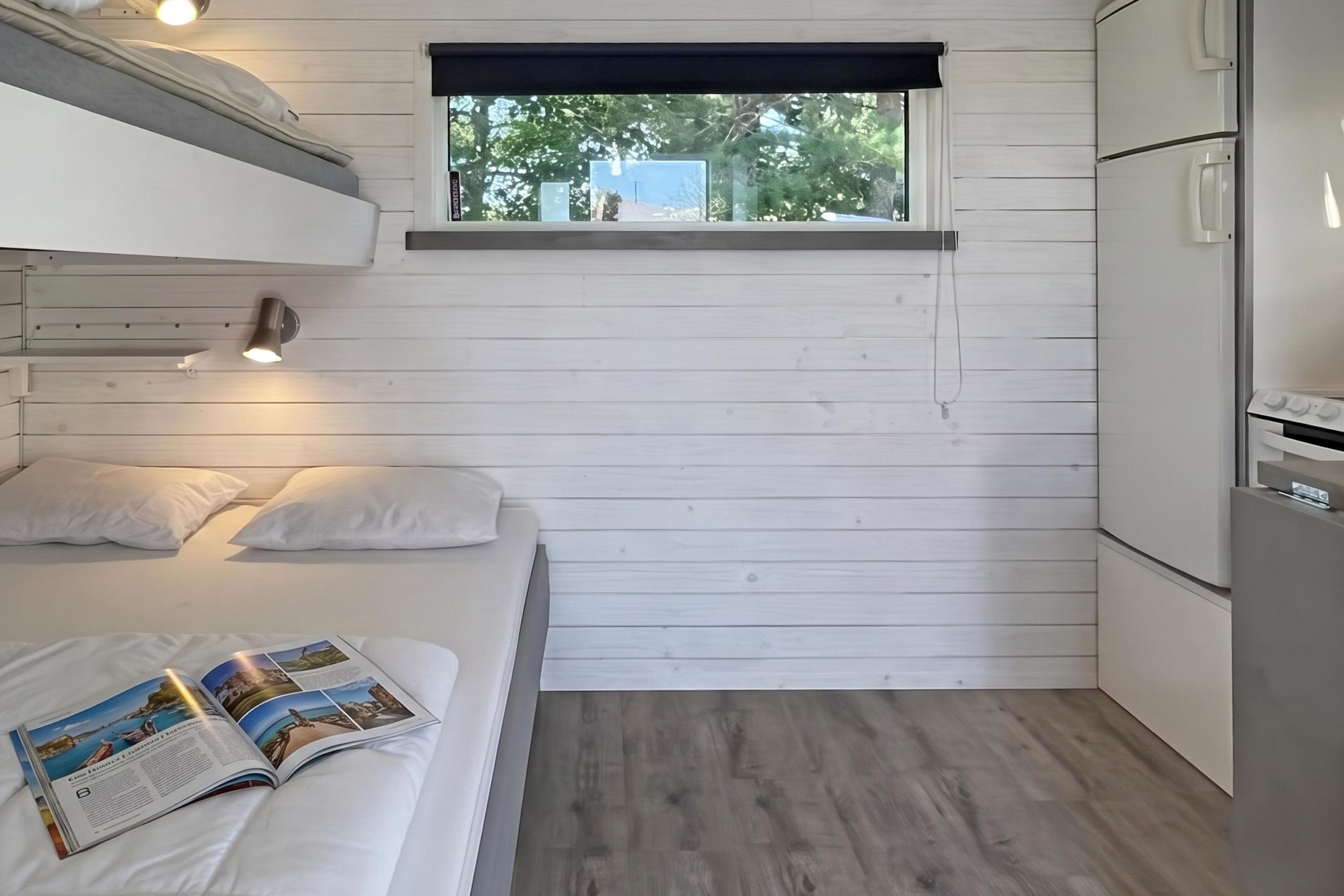 A compact room with a bunk bed, a small window, a kitchenette, and a magazine on the bed.