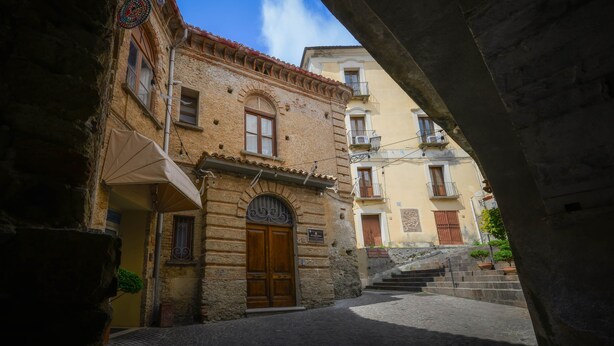 Eine schmale Gasse mit Steinhäusern, einer Holztür und einer Treppe, die zu einer höheren Ebene führt.