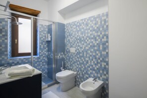 A bathroom with a glass shower enclosure, a toilet, a bidet, and a sink with a towel.