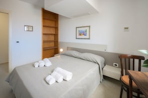 A hotel room with a bed, a wooden shelf, a bedside table, a chair, and a wall-mounted light.