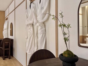 Ein Hotelzimmer mit Holzboden, einem dunklen Holzstuhl, einem Tisch mit Topfpflanze und zwei weißen Bademänteln, die an der Wand hängen.