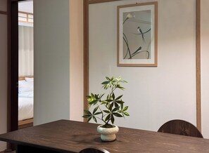 Ein Zimmer mit einem Holztisch, einem Blumentopf, einem gerahmten Bild an der Wand und einem Bett im Hintergrund.