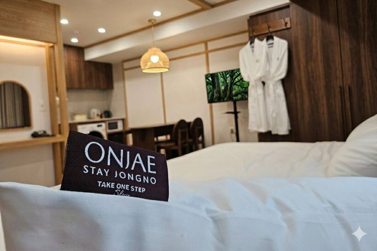 A hotel room with a bed, a sign reading 'ONJAE STAY JONGNO TAKE ONE STEP,' a robe hanging on the wall, and a dining area in the background.